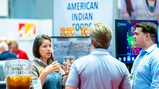 People interacting at an event with a focus on American Indian food, featuring a TV screen and branded logos.