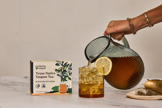 Person pouring tea from a glass pitcher into a glass with lemon slices, next to a box of Texas Native Iced Yaupon Tea.